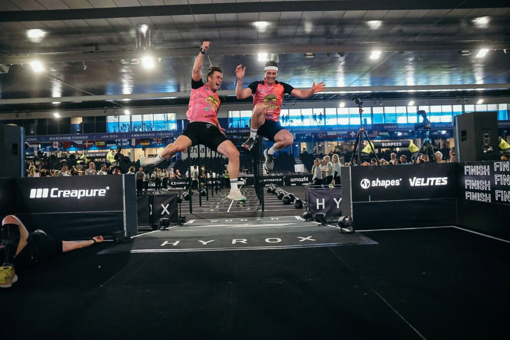 Two men jumping at the finish line of HYROX Men's Doubles.